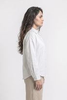 A woman shown in profile wearing a white long-sleeve linen button-up shirt and beige trousers, photographed against a clean white studio background.