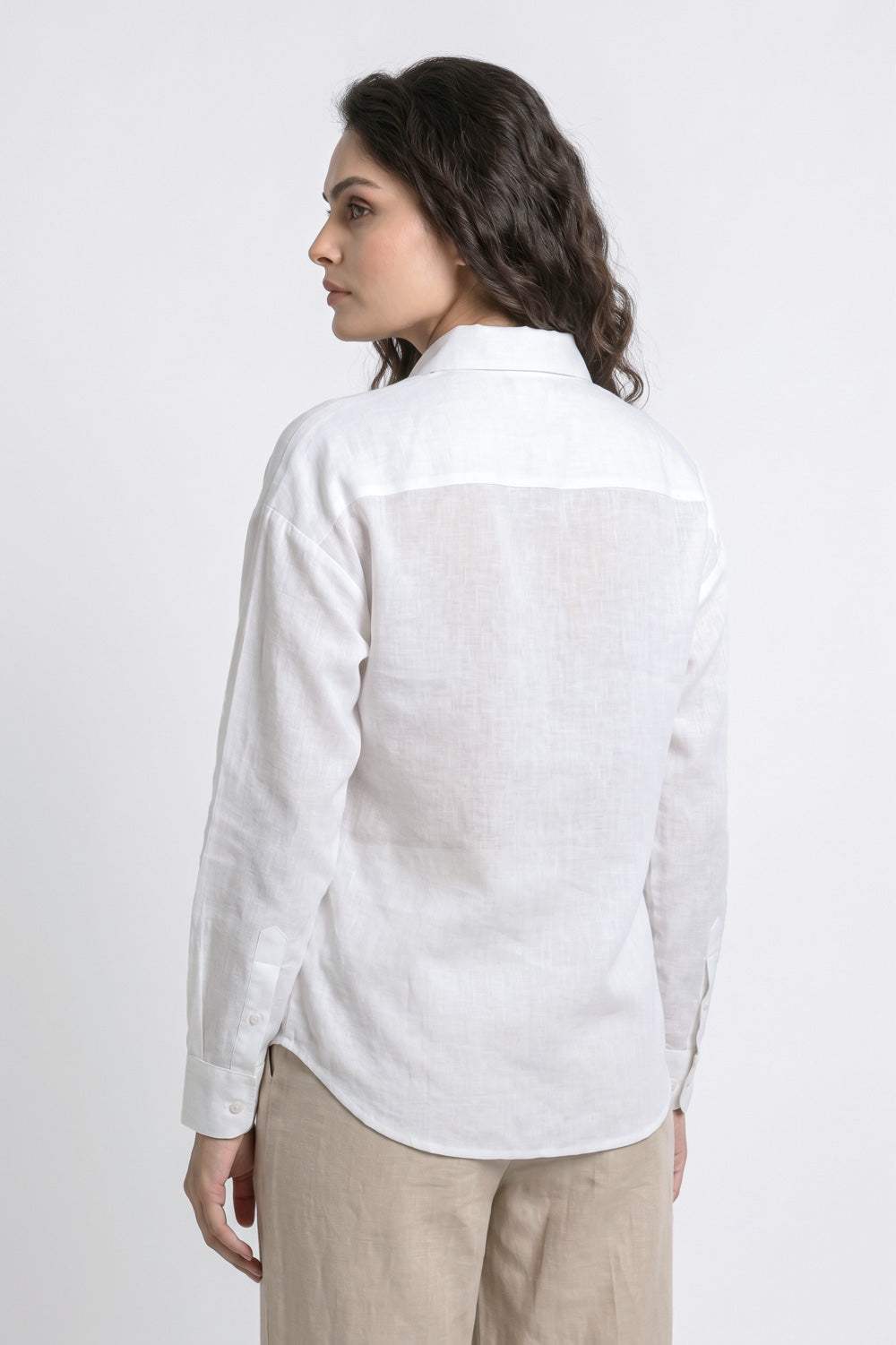 A woman shown from the back wearing a white long-sleeve linen shirt and beige trousers, photographed against a plain light gray studio background.