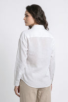 A woman shown from the back wearing a white long-sleeve linen shirt and beige trousers, photographed against a plain light gray studio background.
