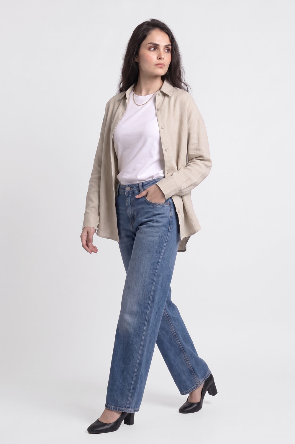 A woman modeling a pair of straight-leg blue jeans, styled with a white t-shirt, a beige button-up linen shirt, and black heels, against a plain light gray studio background.