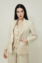 A woman posing against a light gray background, wearing a tailored beige linen three-piece suit. She wears an open single-breasted blazer with notch lapels, a buttoned waistcoat underneath, and matching trousers, keeping one hand resting in her pocket.