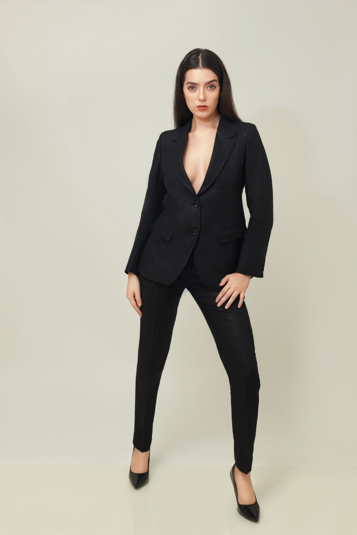 A woman posing against a light gray background, wearing a tailored beige linen three-piece suit. She wears an open single-breasted blazer with notch lapels, a buttoned waistcoat underneath, and matching trousers, keeping one hand resting in her pocket.