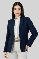 A woman posing against a light gray background, wearing a tailored blue linen blazer. She wears an open single-breasted blazer with notch lapels, a white shirt underneth, and white trousers, with one of her hand on the waist.