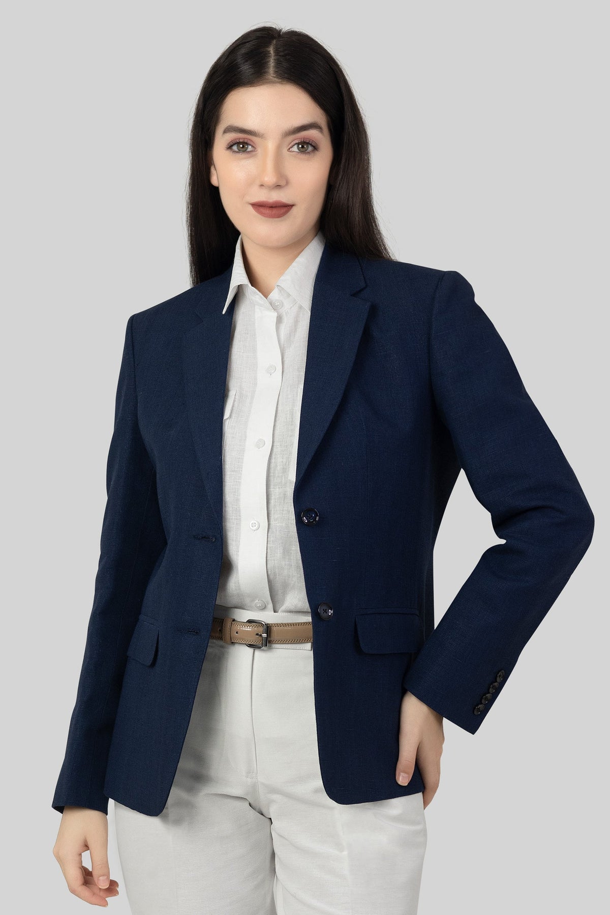 A woman posing against a light gray background, wearing a tailored blue linen blazer. She wears an open single-breasted blazer with notch lapels, a white shirt underneth, and white trousers, with one of her hand on the waist.