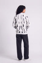 A model shown from the back wearing a white hemp bomber jacket with a black abstract eco-print, paired with black pants and white sneakers, against a light gray studio background.