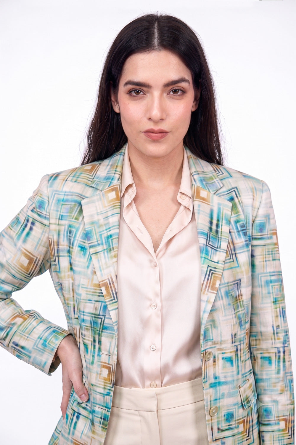 A woman modeling a patterned linen blazer with abstract geometric lines in teal, blue, and gold over a light beige satin shirt and beige trousers, shot against a plain white studio background.