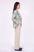 A woman stands in profile wearing a multicolor plaid linen blazer with green, yellow, and blue tones, paired with beige wide-leg trousers and black loafers, against a plain white studio background.