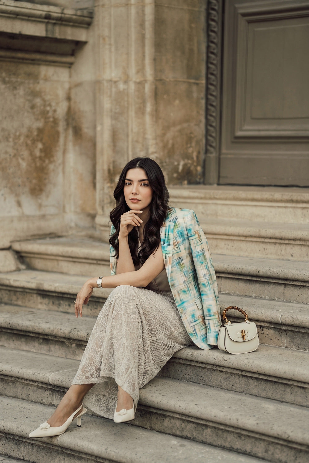 A woman sits on stone steps wearing a beige lace maxi skirt, a light blue and white plaid linen blazer, and white heels, with a small cream handbag beside her.
