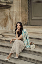 A woman sits on stone steps wearing a beige lace maxi skirt, a light blue and white plaid linen blazer, and white heels, with a small cream handbag beside her.