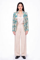 A model stands against a white studio background wearing a long-sleeve patterned linen blazer with teal, green, and beige tones over a pale pink satin shirt and cream wide-leg trousers.