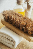 Close-up of a natural coconut coir body brush with a bamboo wood handle, resting on a rolled organic cotton towel beside glass bottles of essential oils — perfect for eco-conscious exfoliation and dry brushing.

