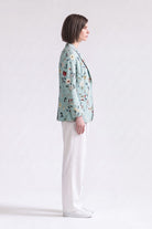 A woman in profile wearing a light blue-green floral linen blazer, white trousers, and white sneakers, standing against a plain white studio background.