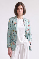 A woman wearing a light sage green floral linen blazer over a white satin top and white trousers, standing against a plain light gray studio background.