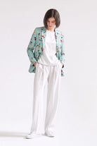 A model stands against a plain white studio background wearing a mint green floral patterned linen blazer over a white t-shirt and white wide-leg trousers.
