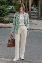 A woman stands on a city street wearing a light mint green floral linen blazer over a white top and cream-colored wide-leg trousers, holding a brown leather handbag.