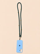 A light blue phone case with a dark green lanyard strap adorned with small gold studs, shown on a solid pale peach background.