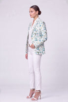 A woman stands in profile wearing a light-colored linen blazer with a blue and green floral pattern, white pants, and white high-heeled sandals against a plain light gray studio background.
