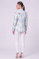 A woman shown from the back wearing a light blue and white floral linen blazer, white pants, and beige high-heeled sandals against a plain light gray studio background.