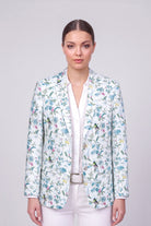 A woman modeling a white linen blazer with a colorful floral and bird pattern, worn over a white collared shirt and white trousers against a light gray studio background.