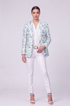 A model stands against a light gray studio background wearing a white button-up shirt, white pants, a white belt, and a light blue and white floral patterned linen blazer, along with white heeled sandals.