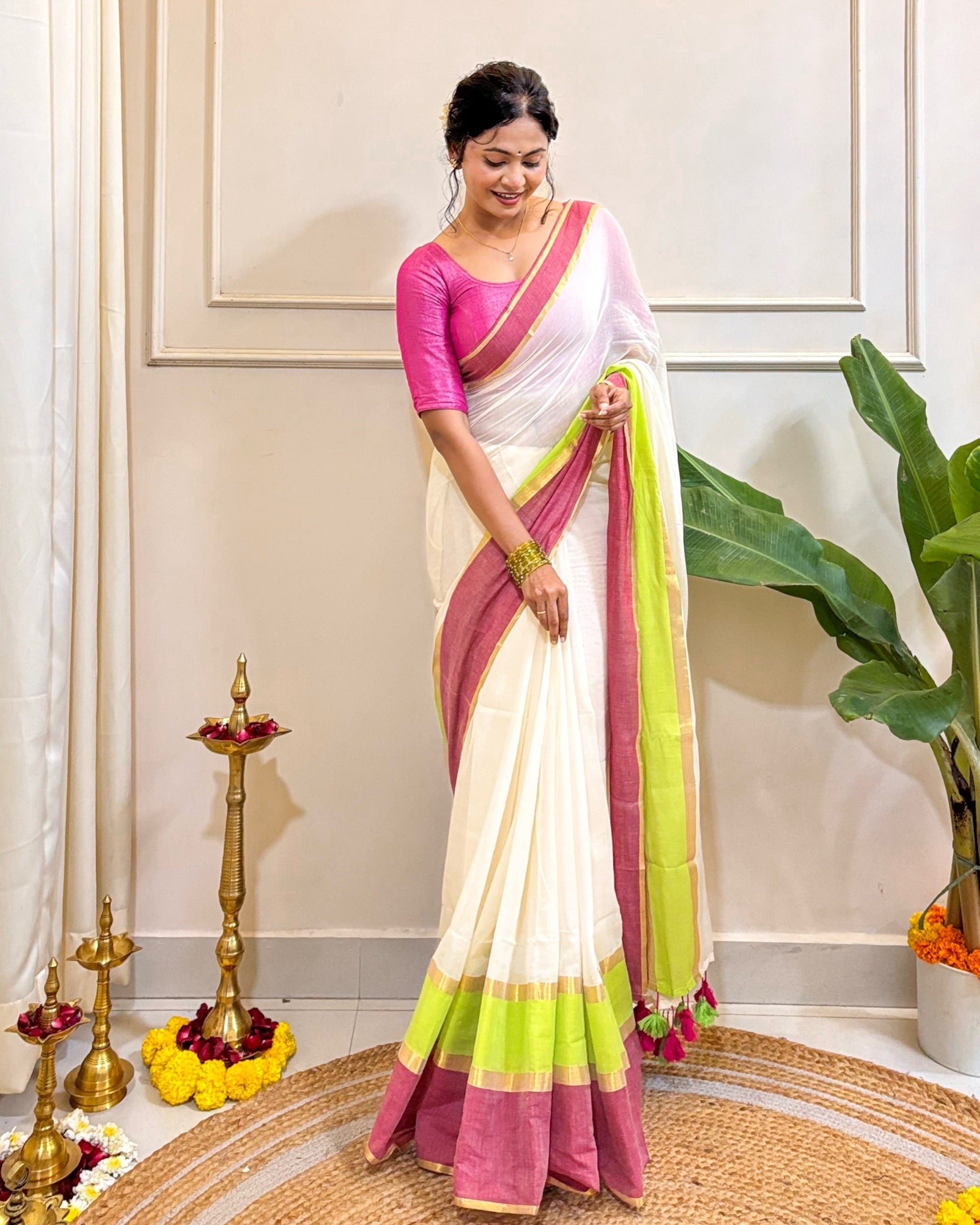 Woman in a white saree with pink and green border standing in a decorated room.