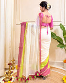 Woman in a white saree with pink and green border standing in a decorated room.