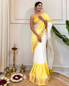 Woman in a yellow and white saree standing indoors with decorative items on the floor.
