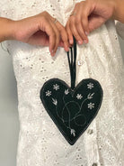 A heart-shaped black fabric bag with metallic thread embroidery, held in hands against a white lace background.
