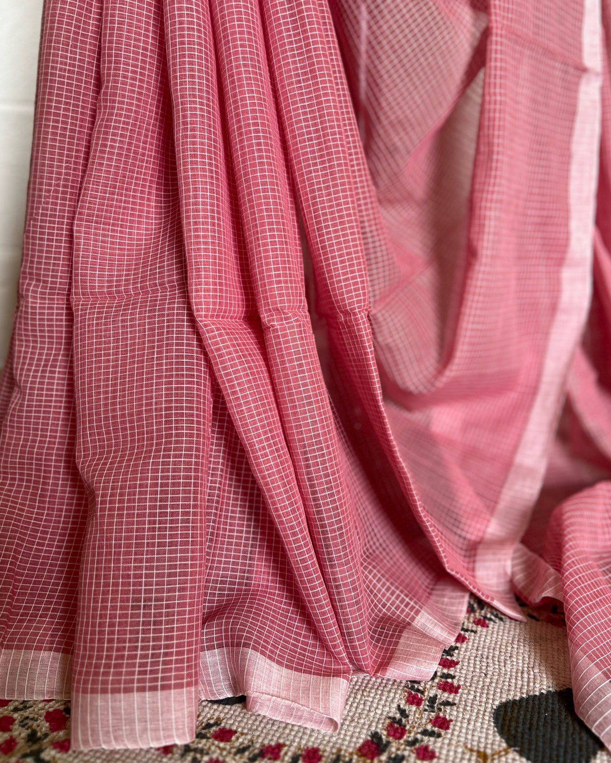 Pink checkered handloom cotton saree draped over a surface with a floral patterned rug underneath.