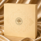 A square cream-colored Royal Bee Brothers gift box with a gold tree of life logo, set against a draped gold satin fabric background.