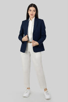 A woman posing against a light gray background, wearing a tailored blue linen blazer. She wears an open single-breasted blazer with notch lapels, a white shirt underneth, and white trousers, holding the blazer with both of her hands.