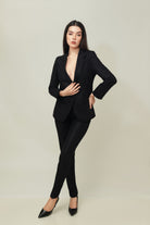 A woman posing against a light gray background, wearing a tailored beige linen three-piece suit. She wears an open single-breasted blazer with notch lapels, a buttoned waistcoat underneath, and matching trousers, keeping one hand at her waist.