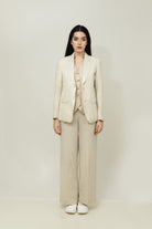 A woman posing against a light gray background, wearing a tailored beige linen three-piece suit. She wears an open single-breasted blazer with notch lapels, a buttoned waistcoat underneath, and matching trousers, keeping her hand resting on sides.
