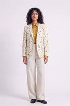 A model stands against a light gray studio background wearing a cream-colored hemp blazer with a yellow and brown eco-print, a mustard yellow collared shirt, cream wide-leg trousers, and black shoes.