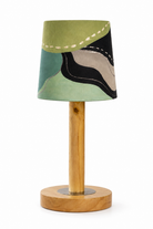 A modern table lamp with a tapered fabric shade featuring an abstract patchwork pattern in sage green, teal, black, and beige, supported by a light natural wood stem and circular wooden base.
