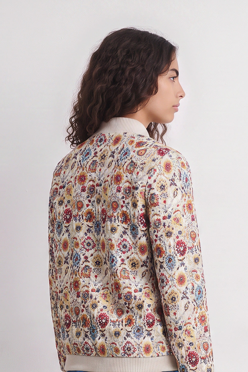 A model shown from the back wearing a cream-colored bomber linen jacket with an all-over dense floral and botanical pattern in red, blue, yellow, and orange.