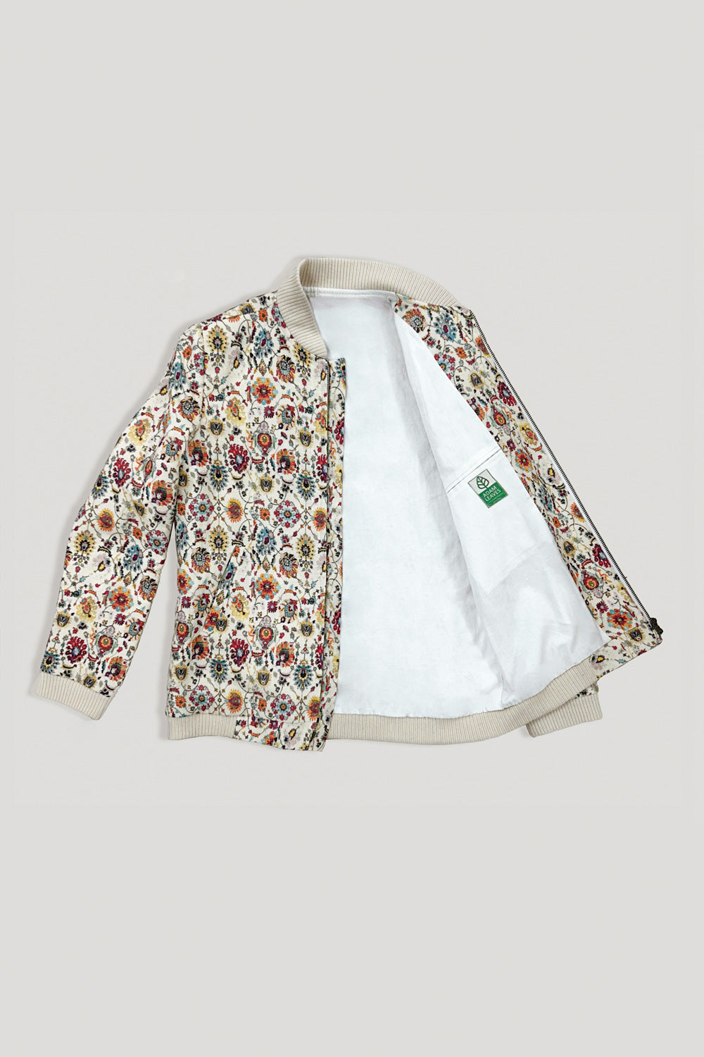A bomber linen jacket with a dense multicolor floral print on a cream base, featuring a white sherpa lining and ribbed beige trim, shown open against a light gray background.