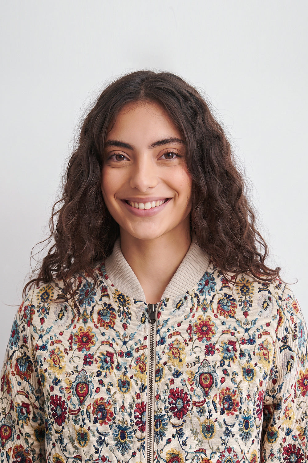 A smiling woman with long, wavy dark brown hair wearing a cream-colored bomber linen jacket with a vibrant, multicolored floral pattern, set against a plain light gray background.