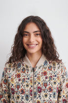 A smiling woman with long, wavy dark brown hair wearing a cream-colored bomber linen jacket with a vibrant, multicolored floral pattern, set against a plain light gray background.