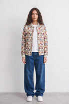 A model stands against a plain light gray wall wearing a cream-colored bomber linen jacket with a dense multicolor floral and botanical print, a white t-shirt, loose-fit blue jeans, and white sneakers.