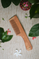 Eco-friendly neem wood comb with handle displayed on a woven mat surrounded by green leaves and tropical flowers, showcasing its smooth texture and zero-waste appeal.