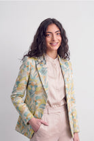 A woman modeling a light beige blouse and matching trousers, styled with a patterned silk blazer featuring abstract yellow, green, and blue floral motifs, against a plain light gray background.
