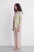A woman stands in profile wearing a light beige top and wide-leg beige trousers, with a pastel tropical print silk blazer in yellow, green, and blue over her shoulders, against a plain white studio background.