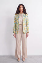 A woman stands against a plain light gray studio wall wearing a beige satin-like blouse and wide-leg beige trousers, with a patterned silk blazer draped over her shoulders. The blazer has a yellow and mint green abstract floral or leaf print.