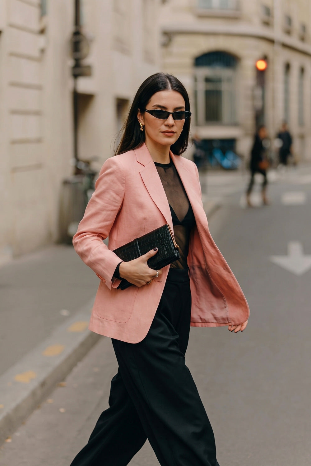A woman walking on a city street wearing a light pink hemp blazer over a sheer black top and black wide-leg trousers, holding a black clutch.