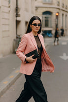 A woman walking on a city street wearing a light pink hemp blazer over a sheer black top and black wide-leg trousers, holding a black clutch.