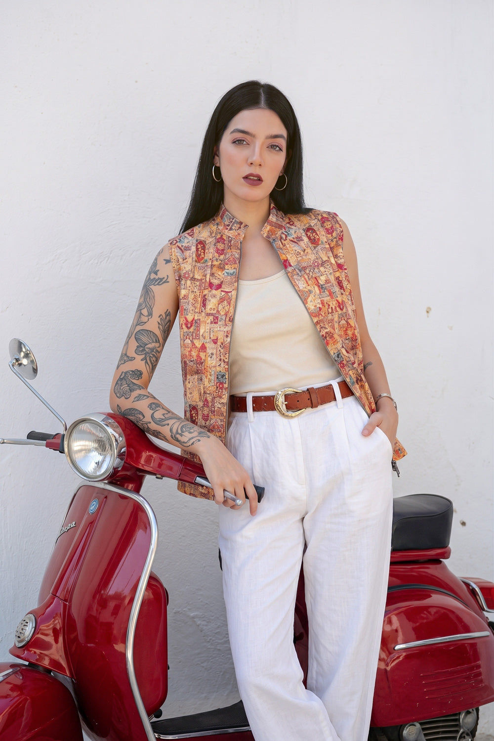 A woman with arm tattoos stands next to a red scooter against a white wall, wearing a sleeveless button-up  linen vest with a colorful floral pattern, a beige tank top, white pants, and a brown belt.