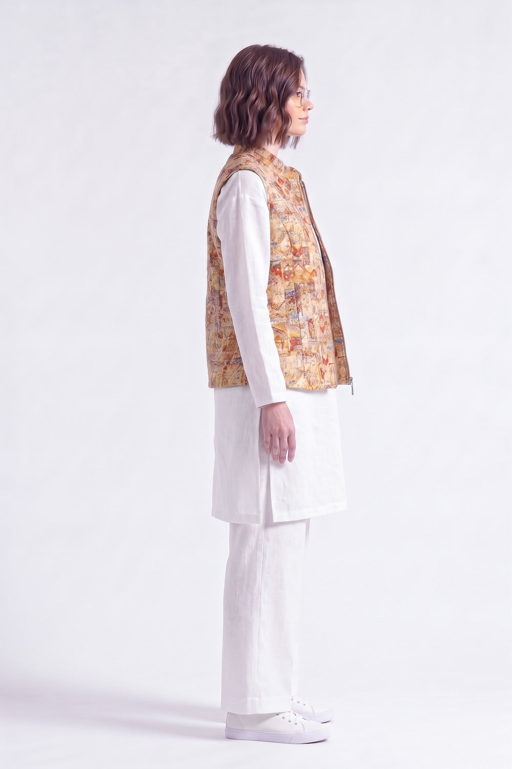 A woman shown in profile wearing a white traditional outfit with a sleeveless patterned  linen vest, photographed against a clean white studio background.