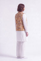 A model shown from the back wearing a white traditional outfit with a patterned mustard and brown linen vest, photographed against a plain white studio background.