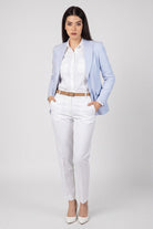 A woman modeling a light blue linen blazer, white shirt, white trousers, and white heels against a light gray studio background.